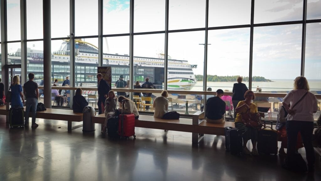 Cruise passengers sitting with suitcases inside a cruise terminal before boarding.