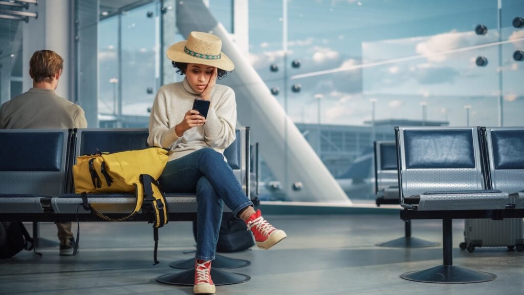 Traveler sitting in an airport looking stressed at her phone, representing common cruise mistakes like flight delays and missed embarkation.