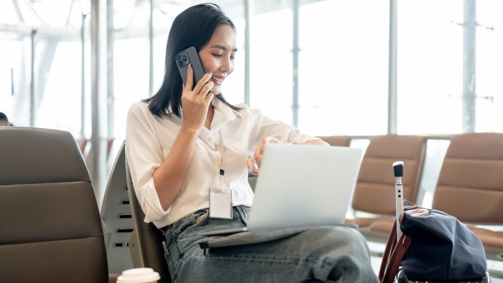 Traveler at airport using laptop and phone while waiting at departure gate with carry-on luggage