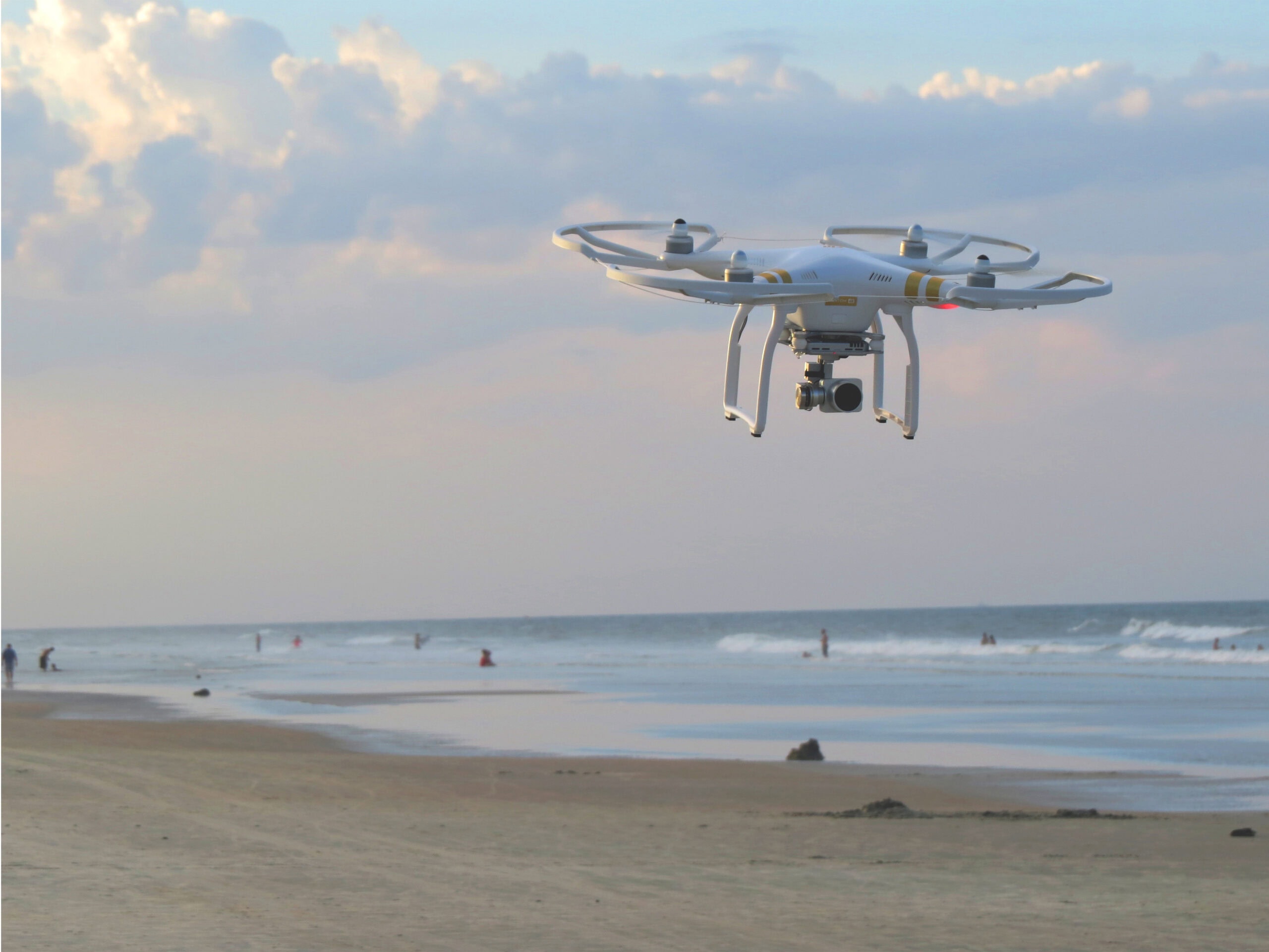 A high end camera drone hovering in the bright blue sky above a tropical sandy beach and turquoise ocean waves.