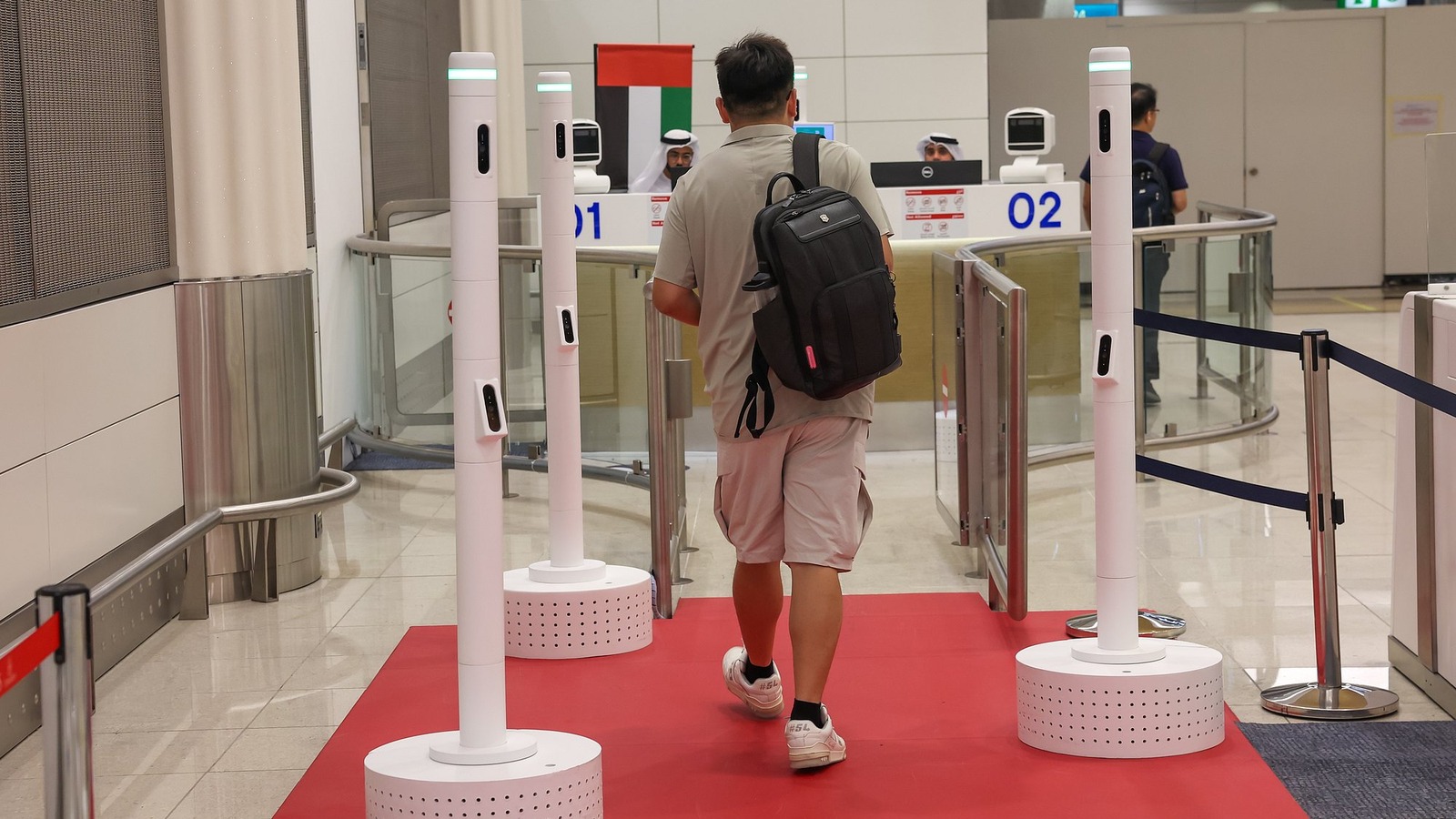 Traveler walking through Dubai International Airport Red Carpet biometric corridor past white sensor poles on a red carpet with UAE flag visible
