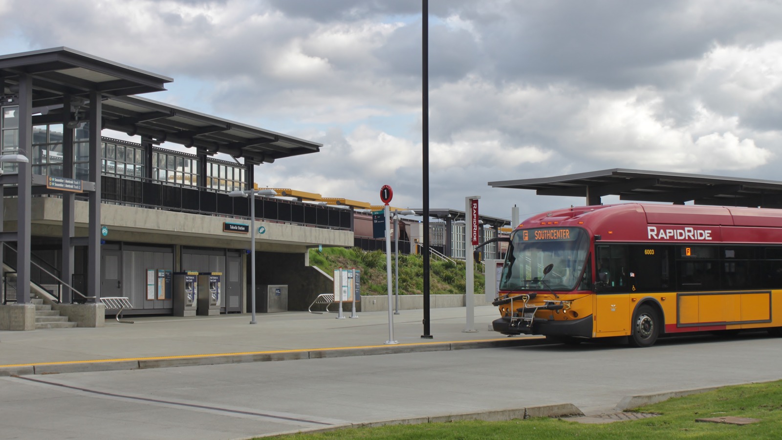 Daylight Tukwila Station entrance with ticket machines and a RapidRide F Line bus at the Longacres Way curb