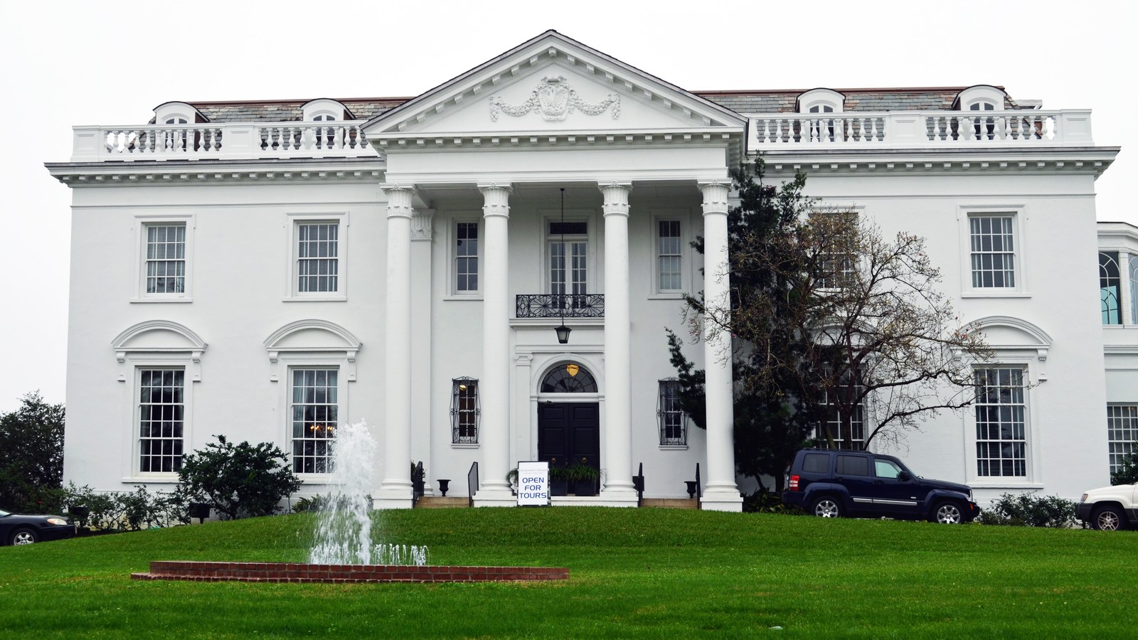 The white columned facade of the Old Governor's Mansion in Baton Rouge behind a lawn and a small fountain under a clear sky