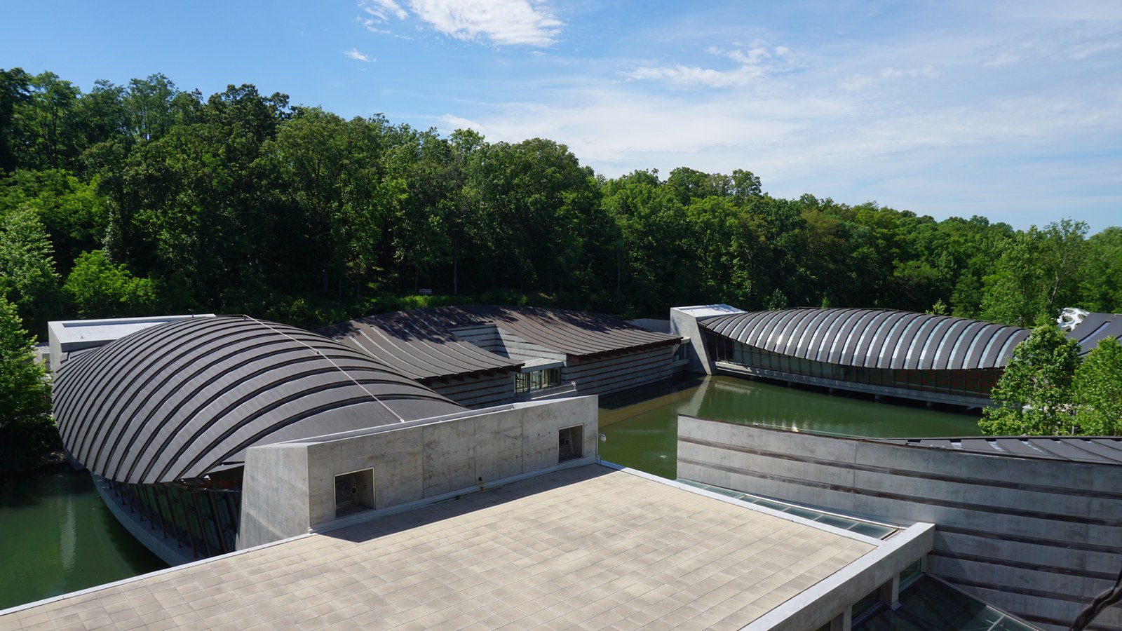 Two curved roofed gallery bridges spanning green ponds at Crystal Bridges in Bentonville, seen from a terrace above with wooded hillside behind