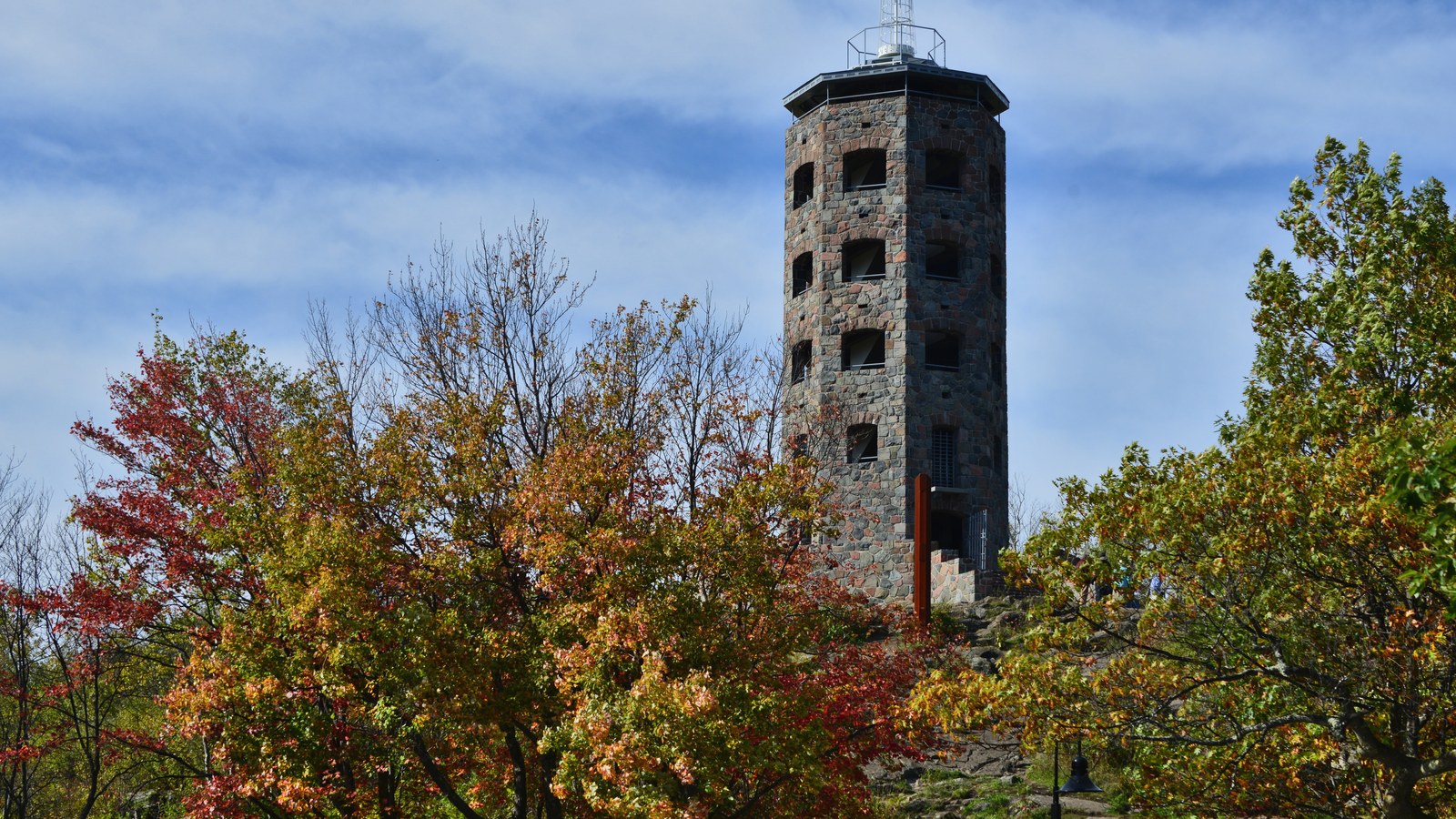The five story blue stone Enger Observation Tower rising above autumn maples on Enger Hill in Duluth, Minnesota, against a blue sky with light cloud