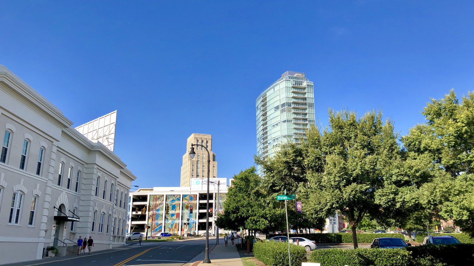A sunlit downtown Durham street with a glass tower and the Hill Building above green trees