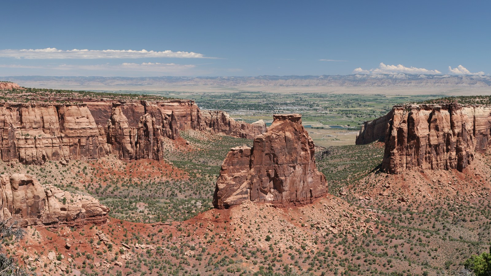 Red sandstone canyon walls and a freestanding rock monolith in Colorado National Monument, with the green Grand Valley and the pale Book Cliffs stretching away behind under a wide sky
