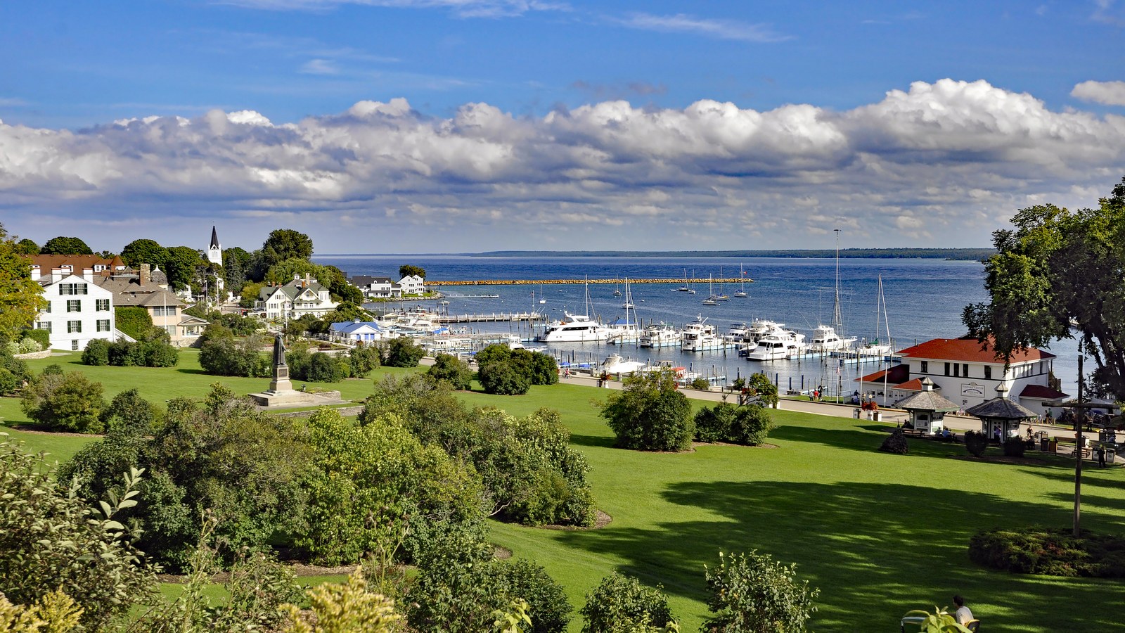 Boats moored in Mackinac Island harbor below the green lawn of Marquette Park and white clapboard buildings, under a bright blue sky