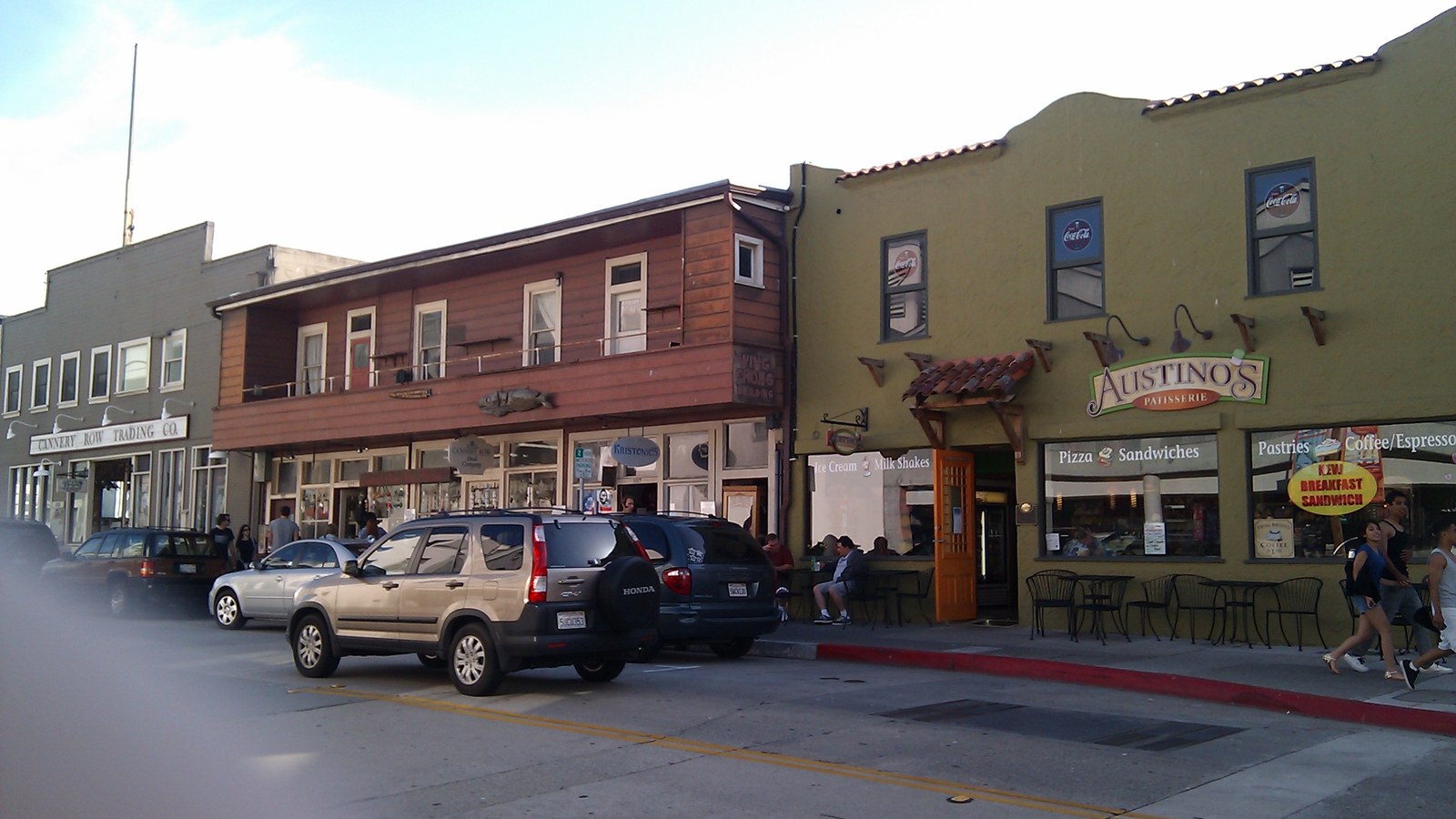 The low wooden and stucco buildings along Cannery Row in Monterey, California, with parked cars and a restaurant awning in bright daylight