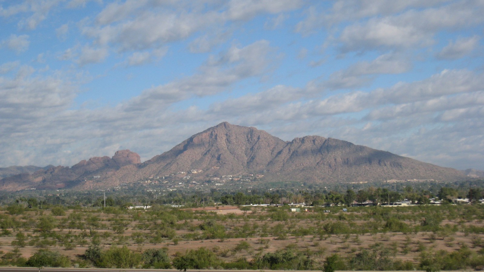 The red rock hump of Camelback Mountain rising above desert scrub and low houses in Phoenix under a broken cloud sky