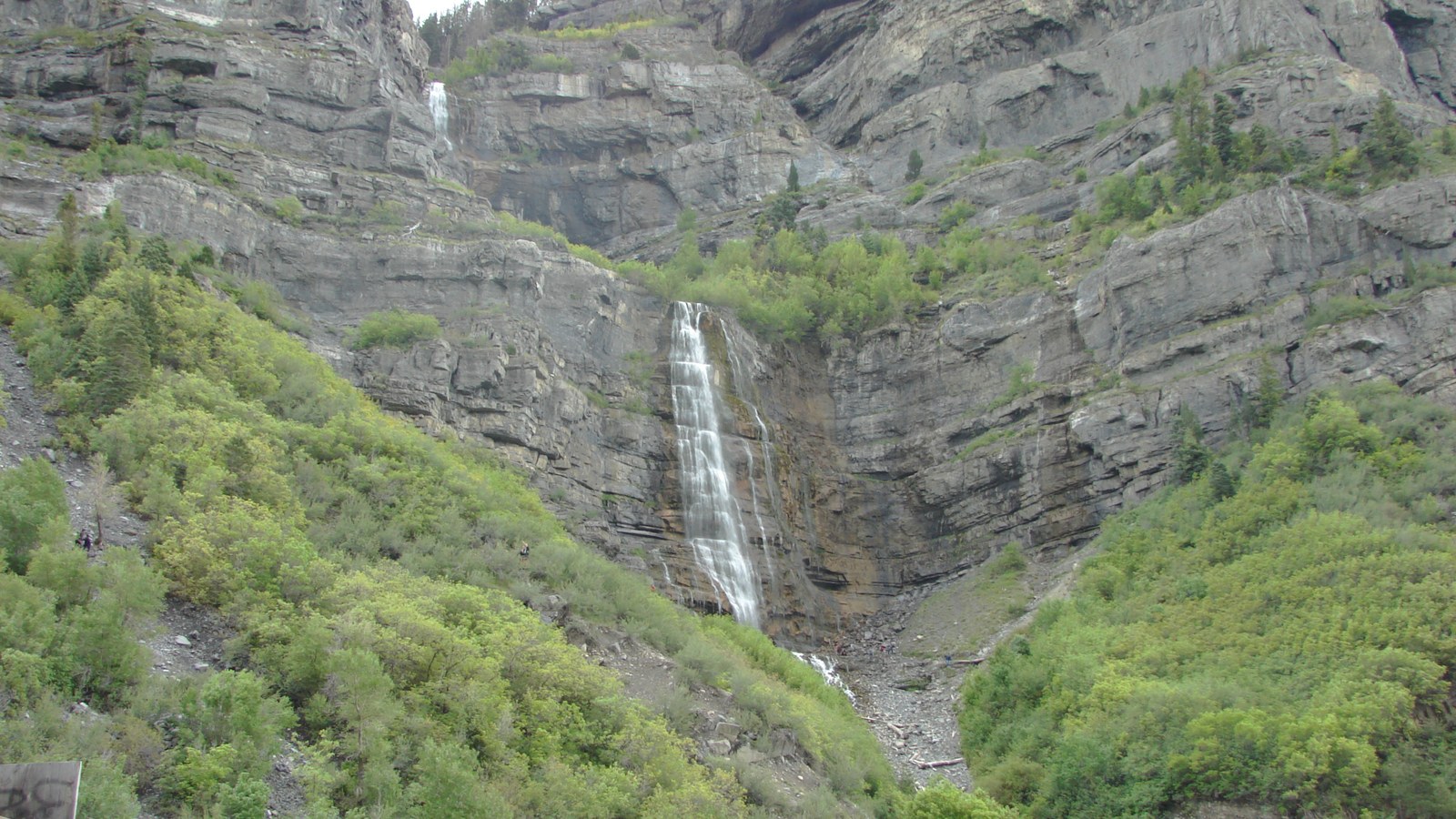 The full double cascade of Bridal Veil Falls dropping down a grey limestone cliff face in Provo Canyon, Utah, with green summer scrub and trees on the slopes either side