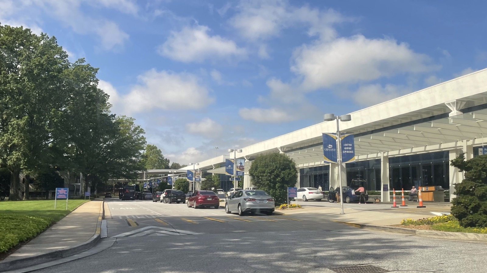 The kerbside drop off and pick up lane outside the terminal at Greenville Spartanburg International Airport, with cars at the kerb under the canopy and blue GSP Airport banners on the light poles