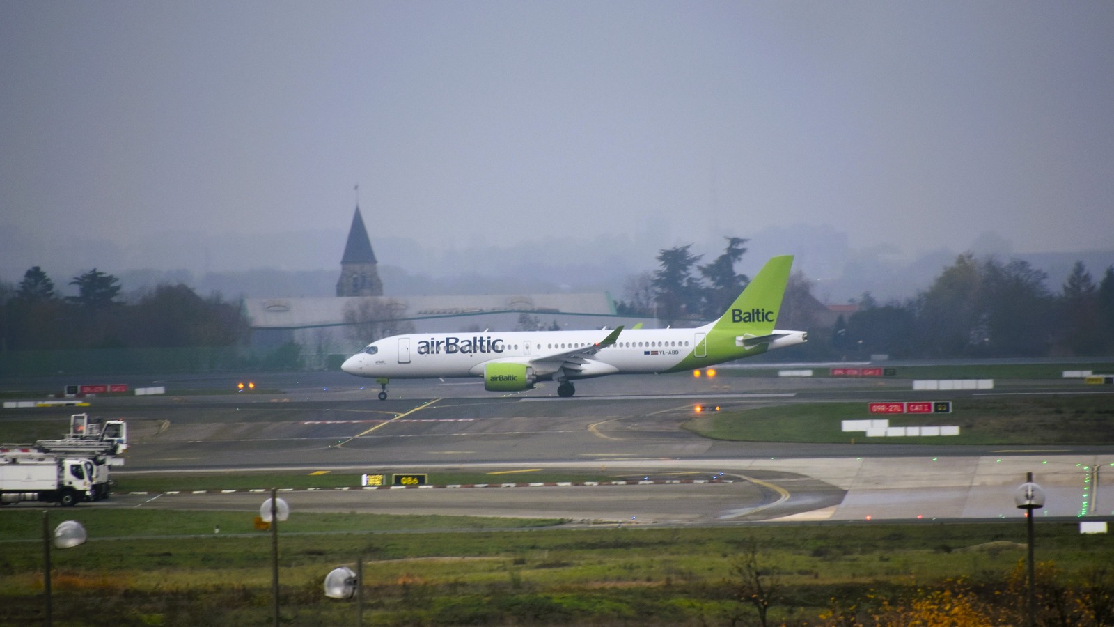An airBaltic Airbus A220 with a green tail lands on a runway in the airline's current livery.