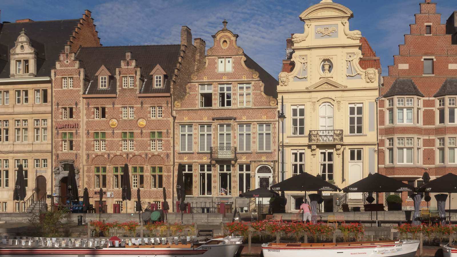 Historic guild houses with stepped and ornate gables along the Korenlei waterfront in Ghent, with canal boats and red flowers in front.