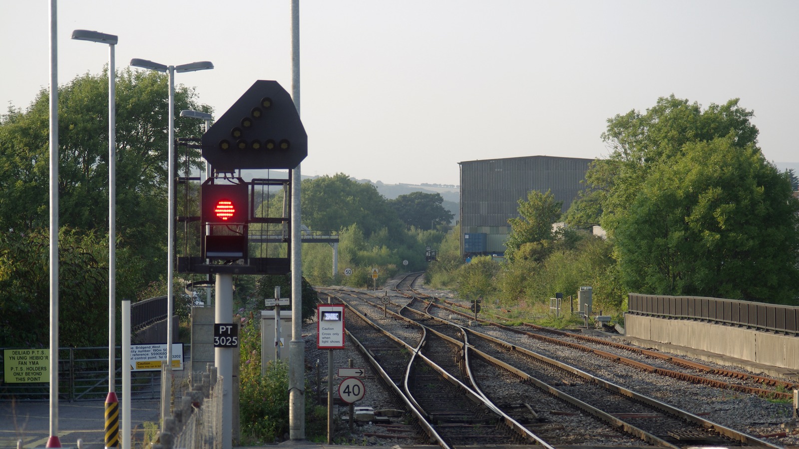 The railway east of Bridgend station looking towards the junction, with a colour light signal at red on a gantry, sets of points and crossovers in the foreground, a bilingual Welsh and English permit to work sign beside the track, and speed restriction signs along the line