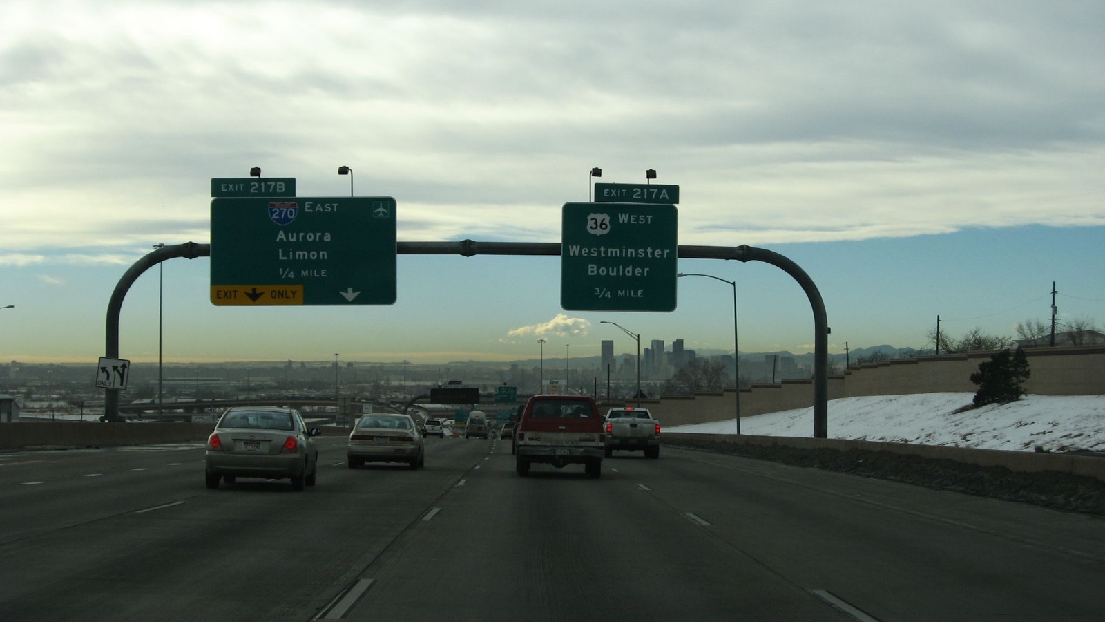 The approach to the Interstate 25 interchange in Denver with an overhead sign gantry carrying Exit 217B for Interstate 270 east to Aurora and Limon marked exit only, and Exit 217A for US 36 west to Westminster and Boulder, with traffic on the carriageway and the Denver skyline and Front Range beyond