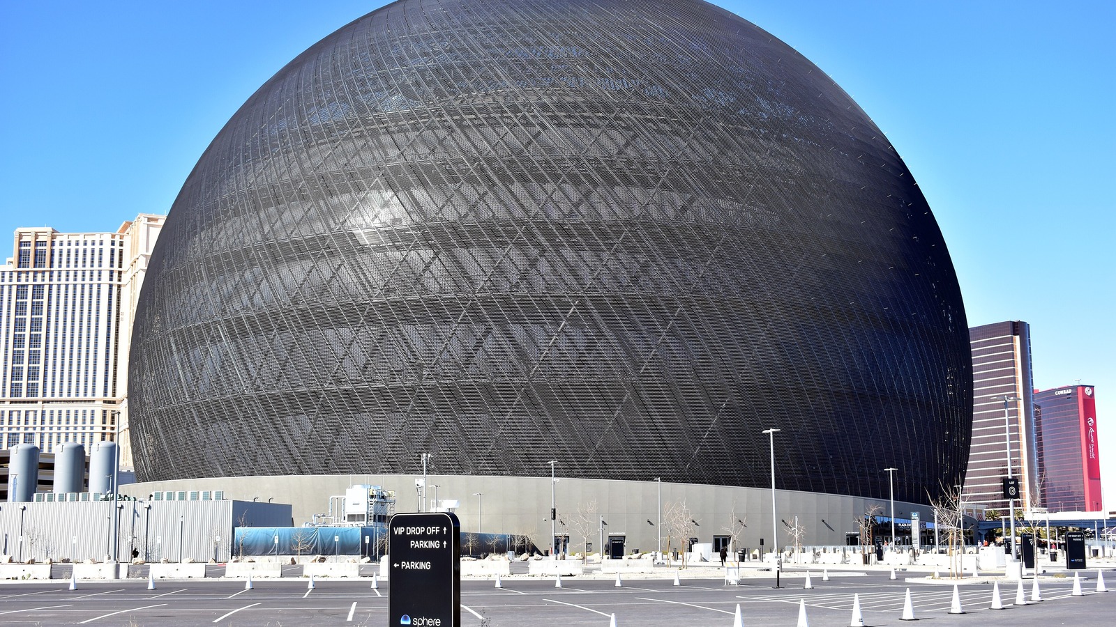 The Sphere in Las Vegas in daylight with its exosphere screens dark, seen across its parking lot with a VIP drop off sign in the foreground and Strip hotel towers behind