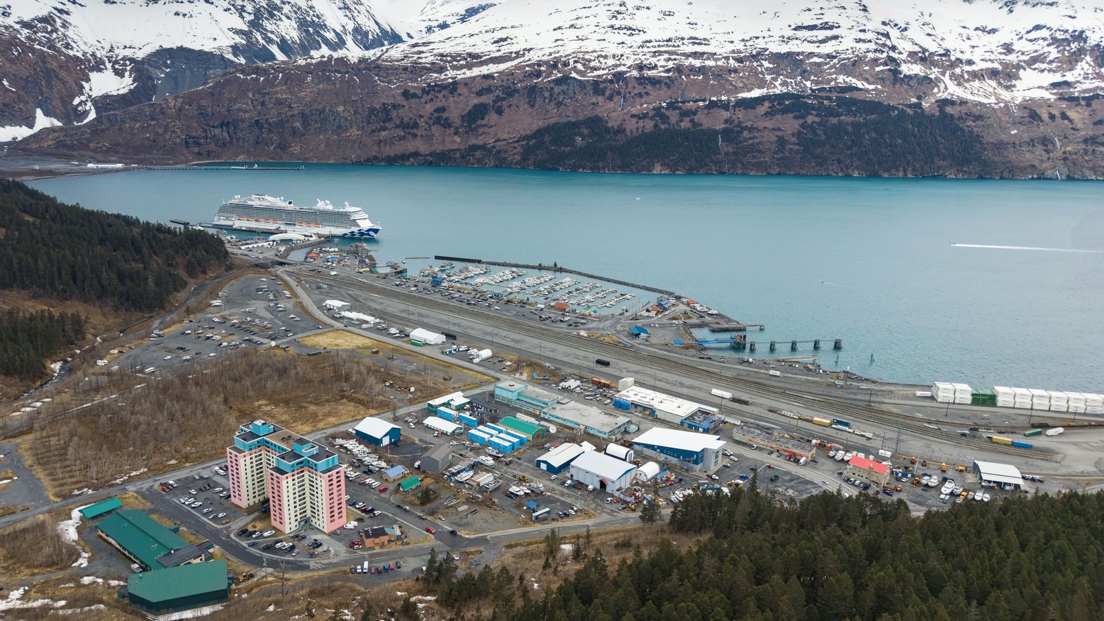Aerial view of Whittier, Alaska showing the cruise dock and a berthed cruise ship, the small boat harbour, the rail yard and Prince William Sound below snow covered mountains