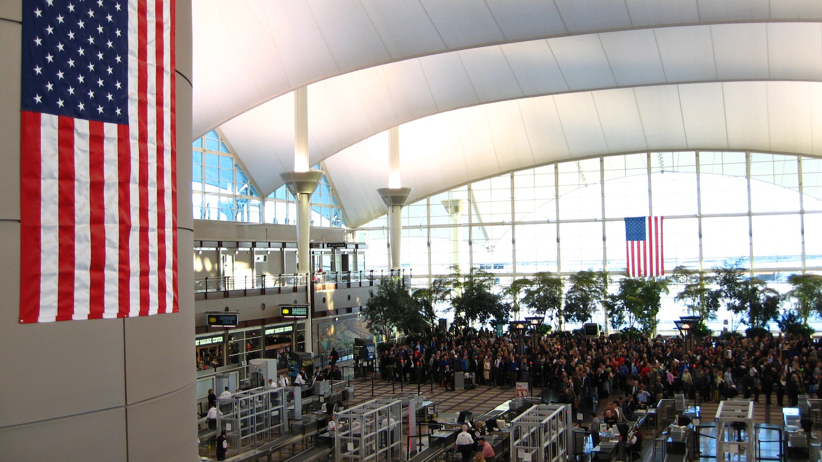 Large crowds queuing for security screening beneath the white tented roof of the main terminal at Denver International Airport, with screening lanes and a United States flag in the foreground.