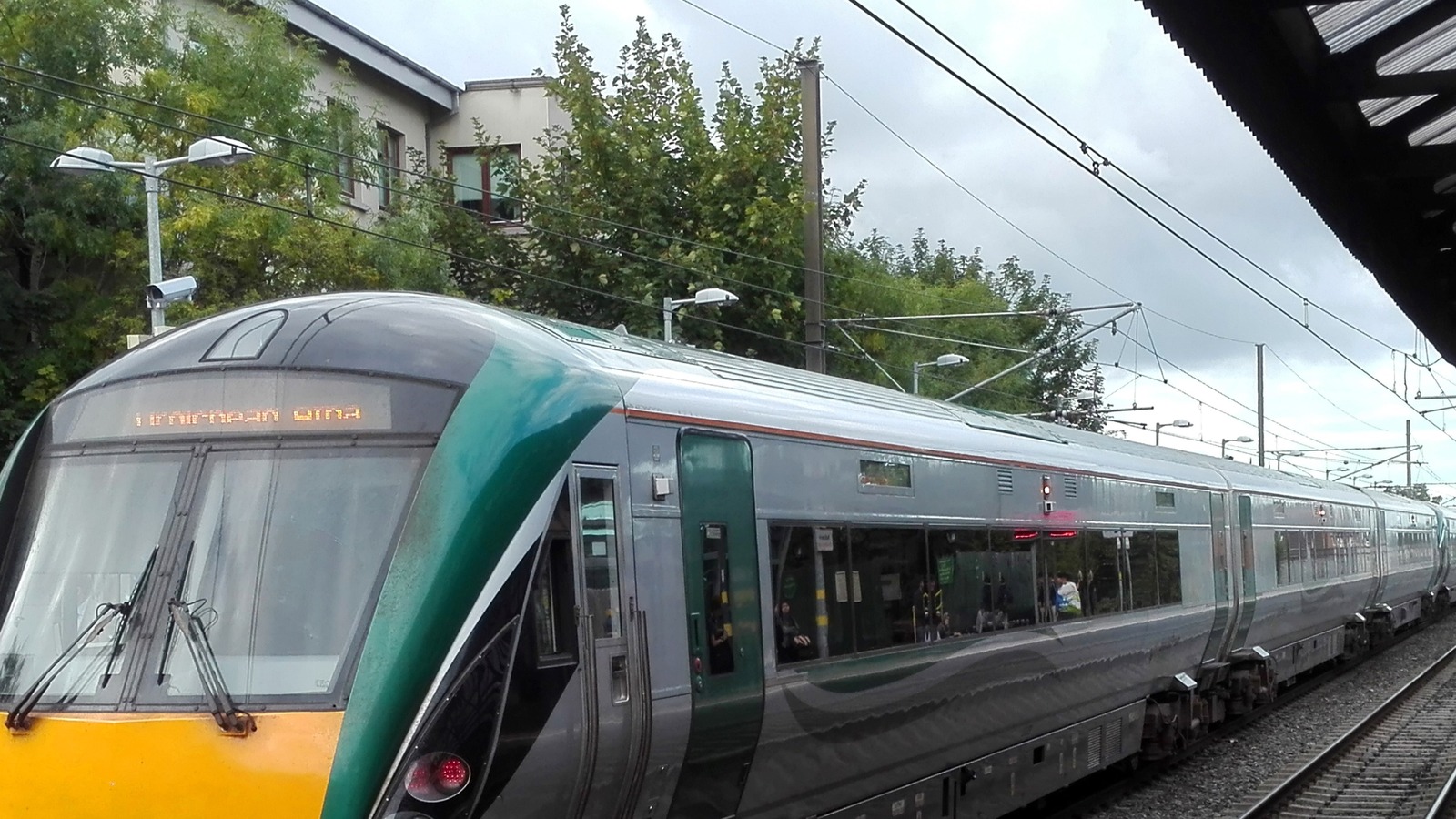 An Iarnrod Eireann InterCity railcar in green and silver livery standing at a station platform with passengers visible through the windows, overhead line equipment above and an illuminated destination display on the cab front