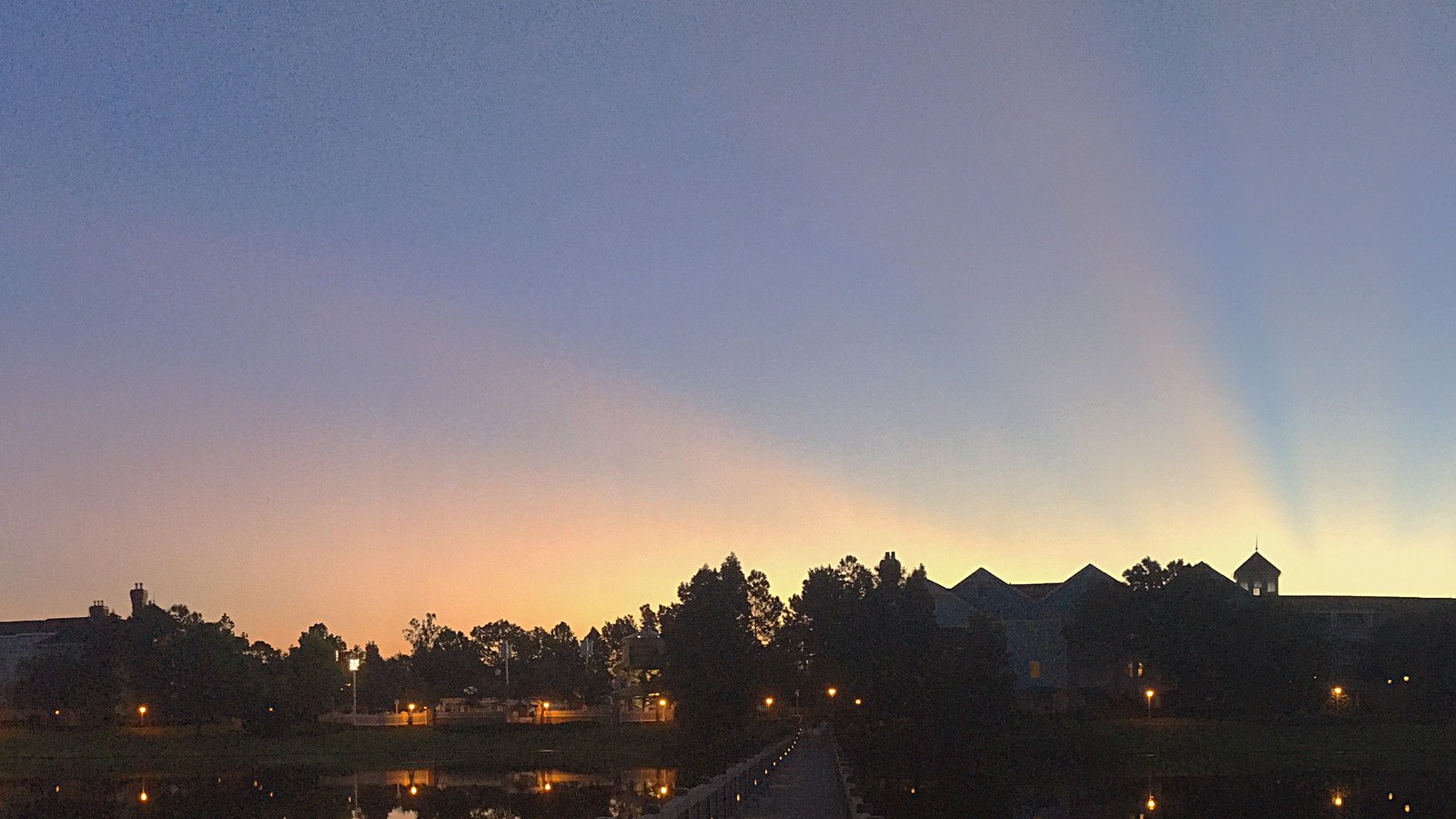 Disney's Saratoga Springs Resort at dusk seen across the lake, with the resort buildings in silhouette against an orange sky and lamps along the walkway