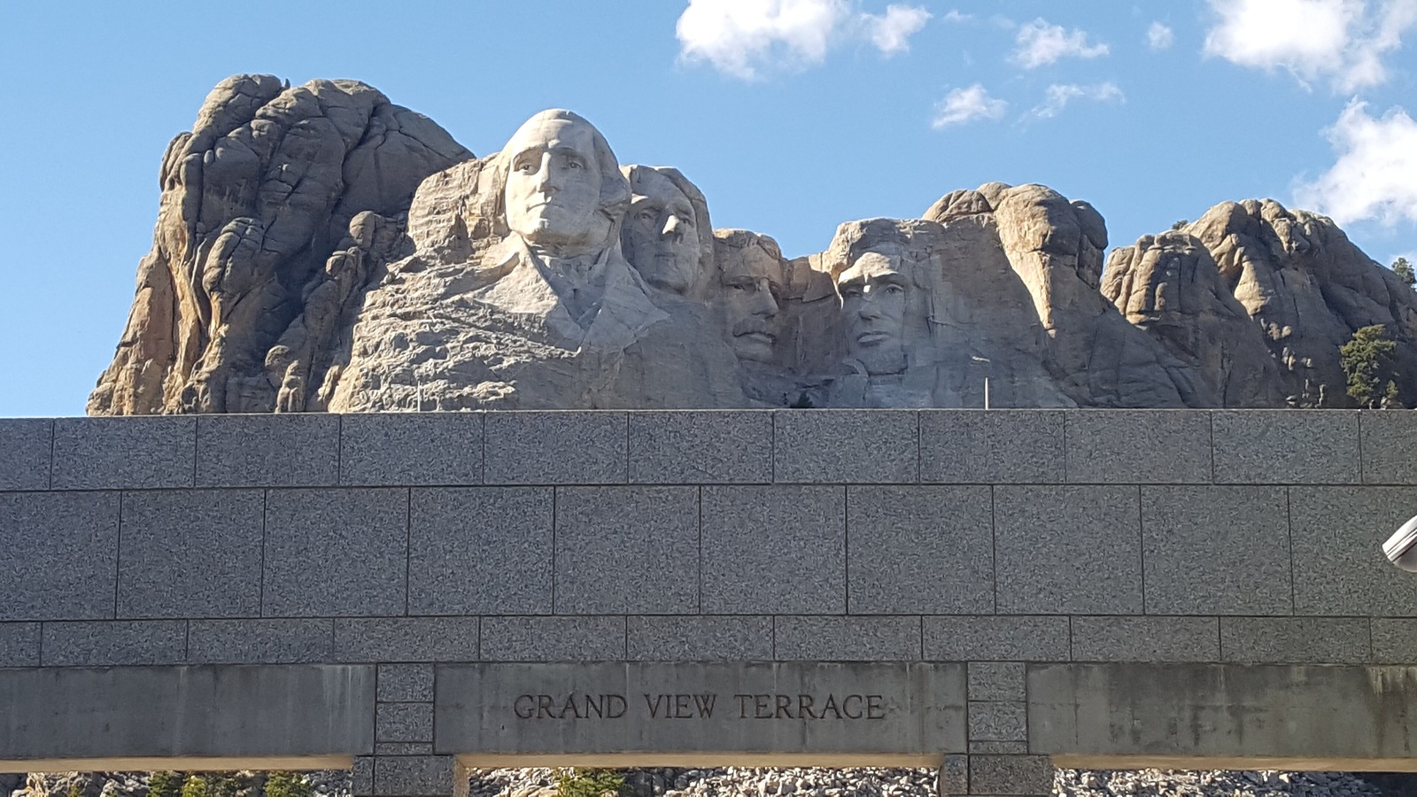 The four carved presidents of Mount Rushmore in afternoon light above the granite wall of the Grand View Terrace, with the terrace name cut into the stone in the foreground.
