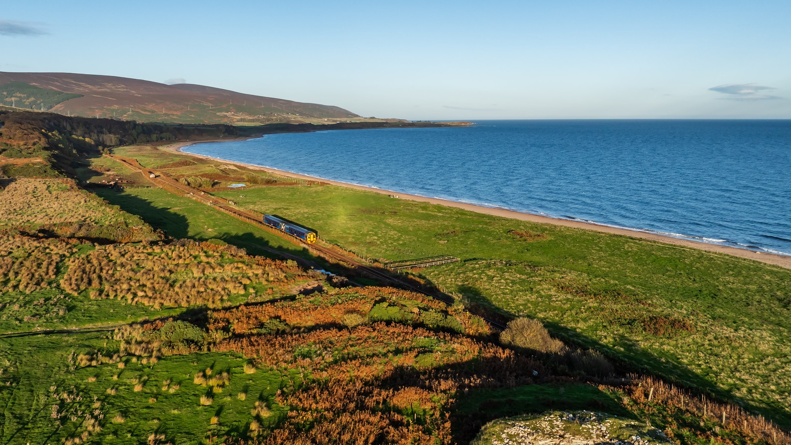 A two car ScotRail Class 158 in Saltire livery running along the single track Far North Line between Brora and Helmsdale, with the coast on one side and autumn bracken turning orange across the hillside