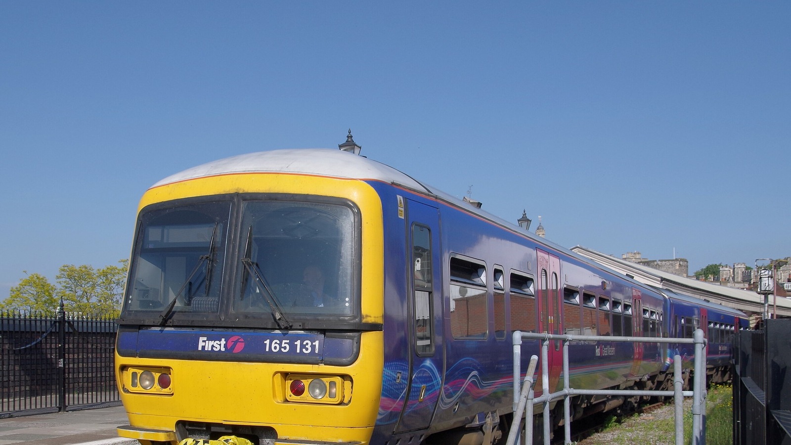 A two car diesel unit in an older purple and yellow livery standing at the single platform of Windsor and Eton Central station under a clear blue sky, with the station canopy running along the right of the train, iron railings to the left and the stone towers of Windsor Castle rising above the rooft