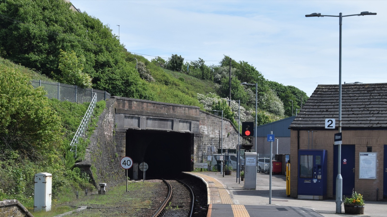 The tunnel portal south of Whitehaven station seen along the single track from the platform end, with a colour light signal at red, a 40 speed restriction board, a token cabinet on the platform and the station buildings and platform 2 sign to the right