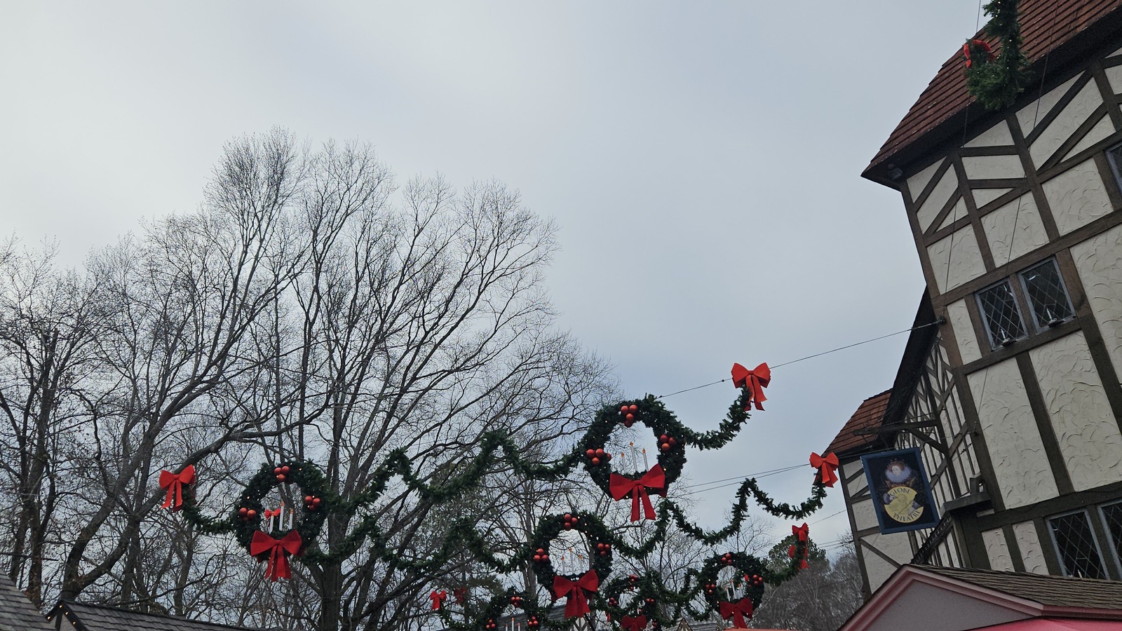 Christmas garlands, wreaths and red bows strung on wires above the England hamlet at Busch Gardens Williamsburg, with a Tudor style timbered building and the Globe Theatre sign on the right under an overcast winter sky