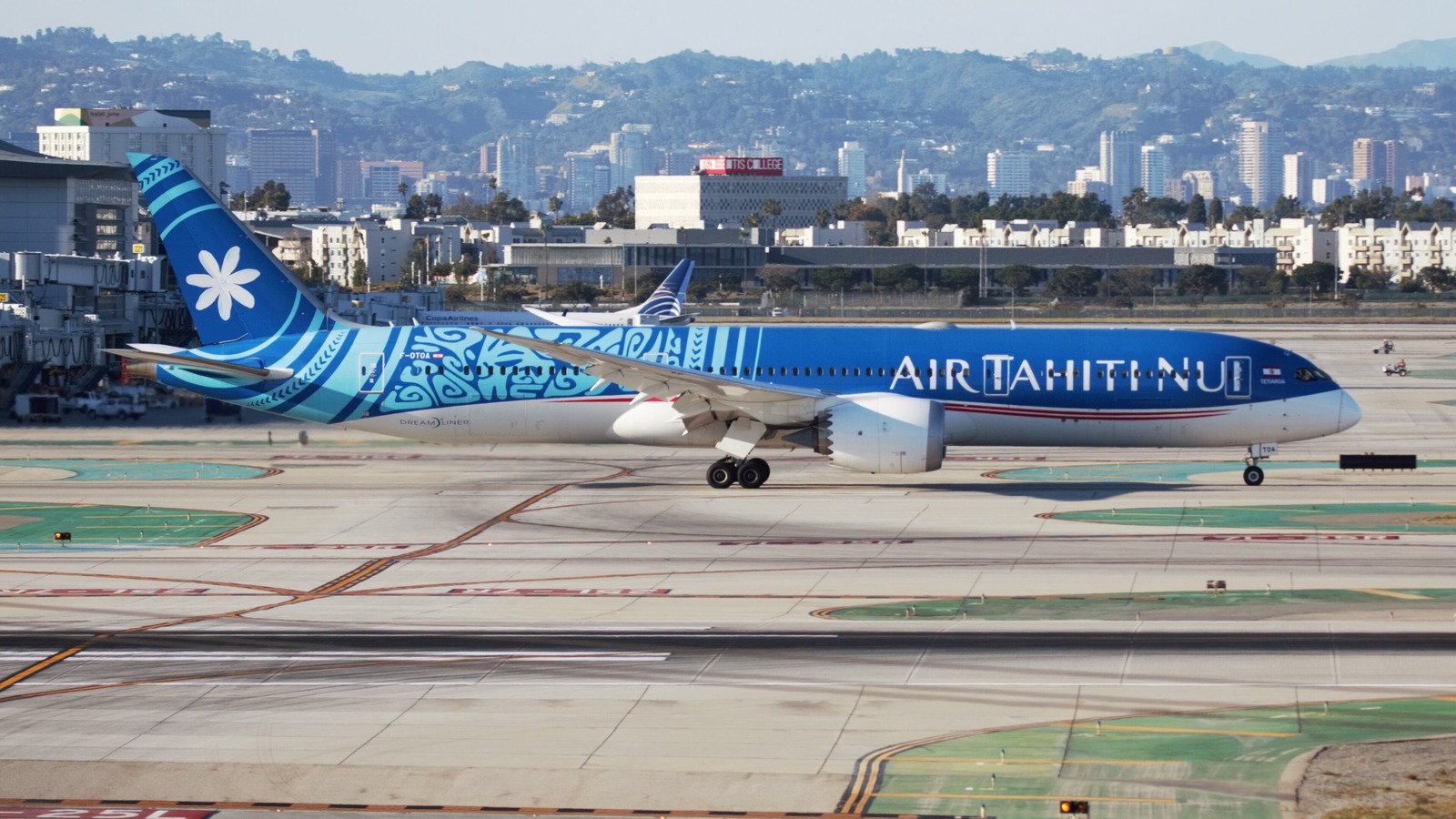 An Air Tahiti Nui Boeing 787-9 in blue Polynesian livery taxiing at Los Angeles International Airport with the city and hills behind.