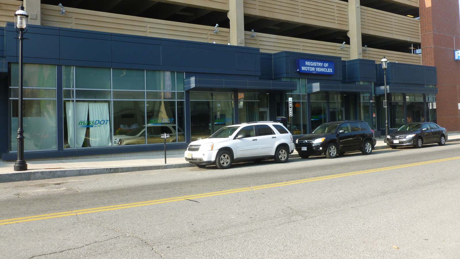 The street front of a Registry of Motor Vehicles office in Lowell, Massachusetts, with its sign above glass doors and cars parked at the curb.
