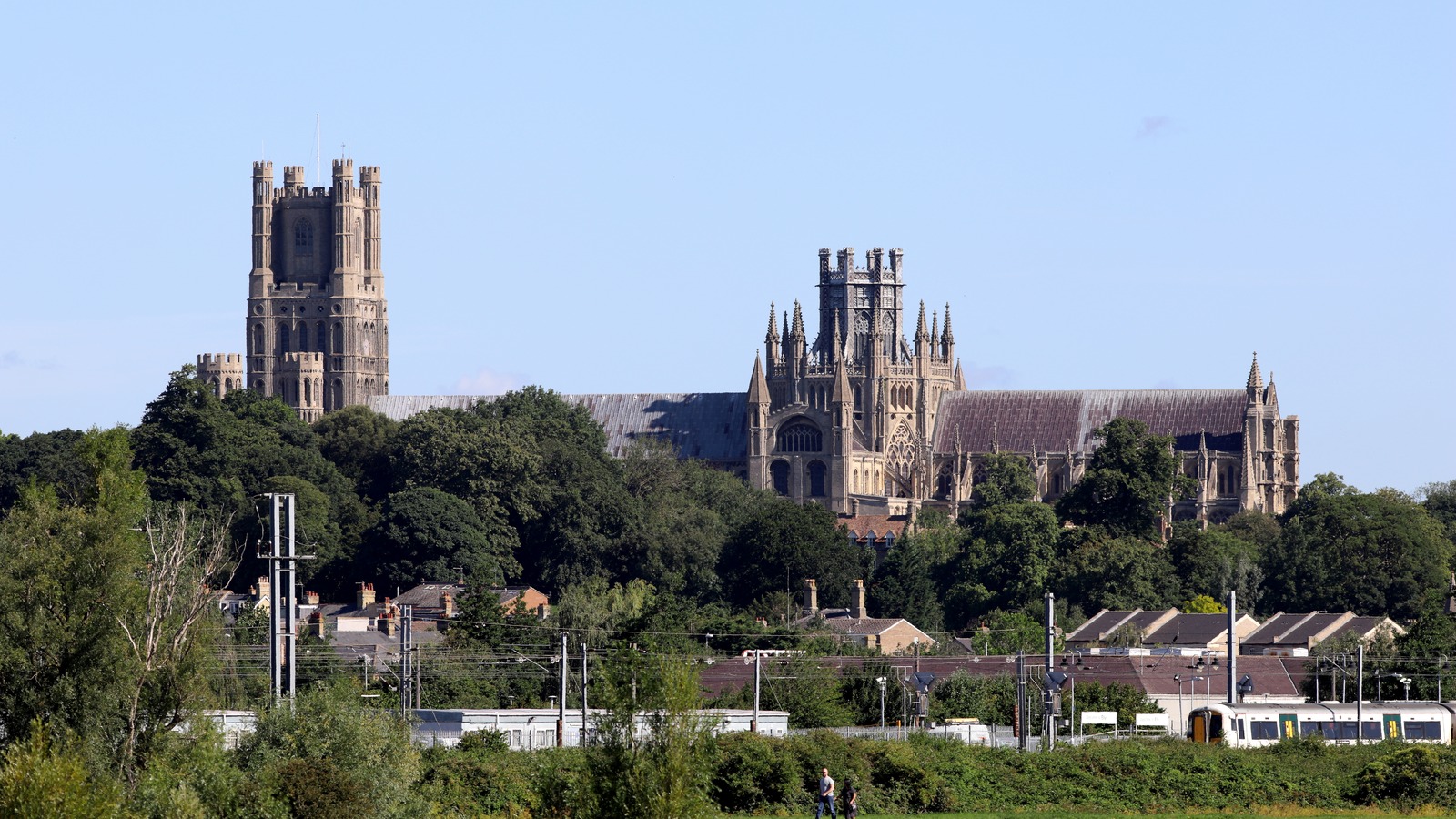 Ely Cathedral rising above trees and rooftops, with the railway, overhead line masts and a passenger train in the foreground