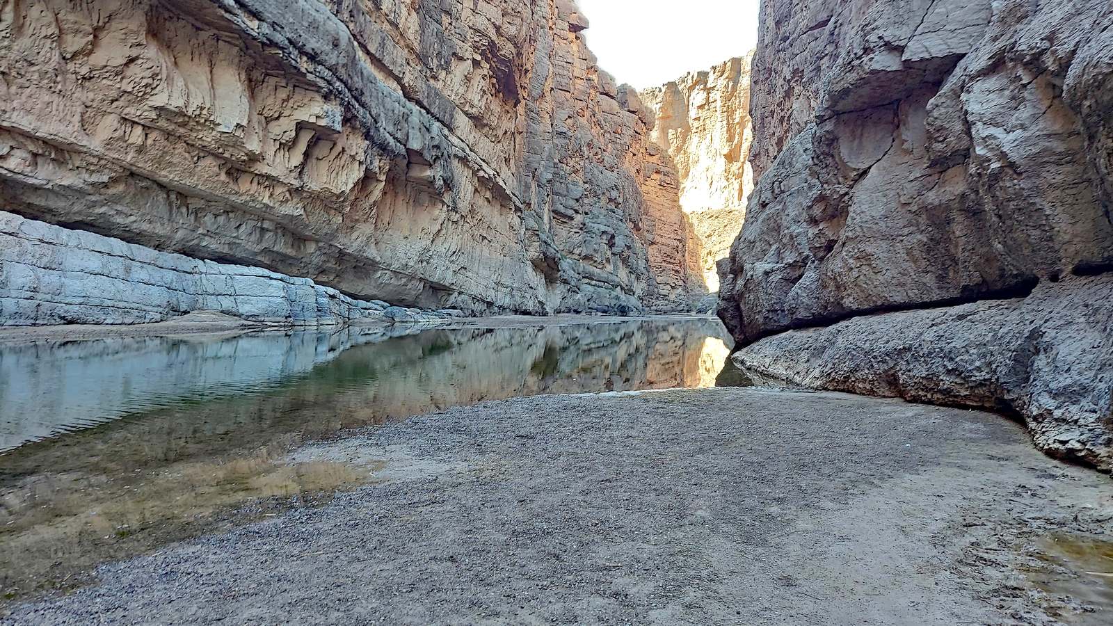 Santa Elena Canyon in Big Bend National Park, its walls rising sheer on both sides of the Rio Grande, with a wide gravel bar in the foreground where the bank meets the water.