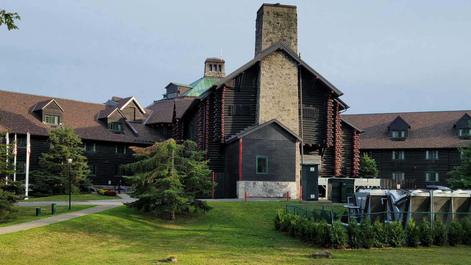 The dark-stained log main lodge of Château Montebello in Quebec, with its tall stone chimney, gabled roofs and lawn, photographed in July 2022.