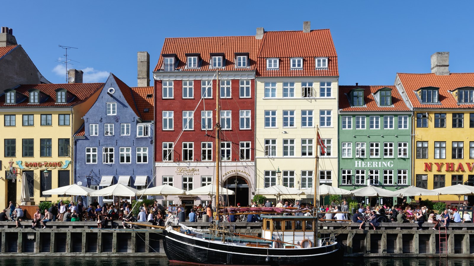 The coloured townhouses along Nyhavn in Copenhagen on a clear afternoon in June 2022, with a wooden sailing boat moored at the quay and people at the cafe tables along the waterfront