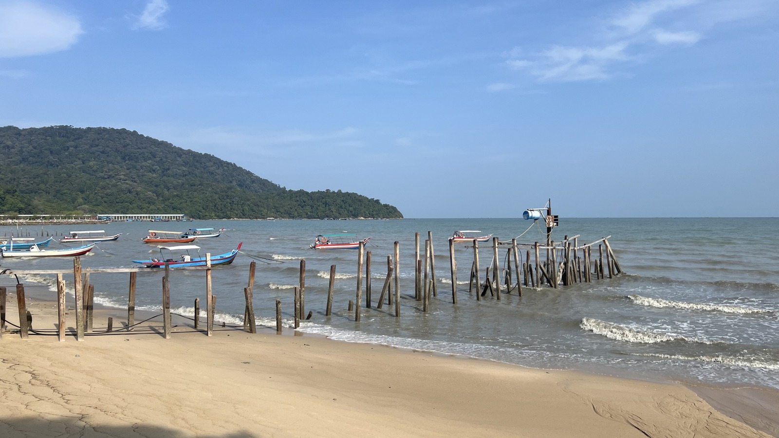Daytime view of the Teluk Bahang shoreline in Penang with fishing boats, wooden jetty pilings, sandy beach and a forested headland.