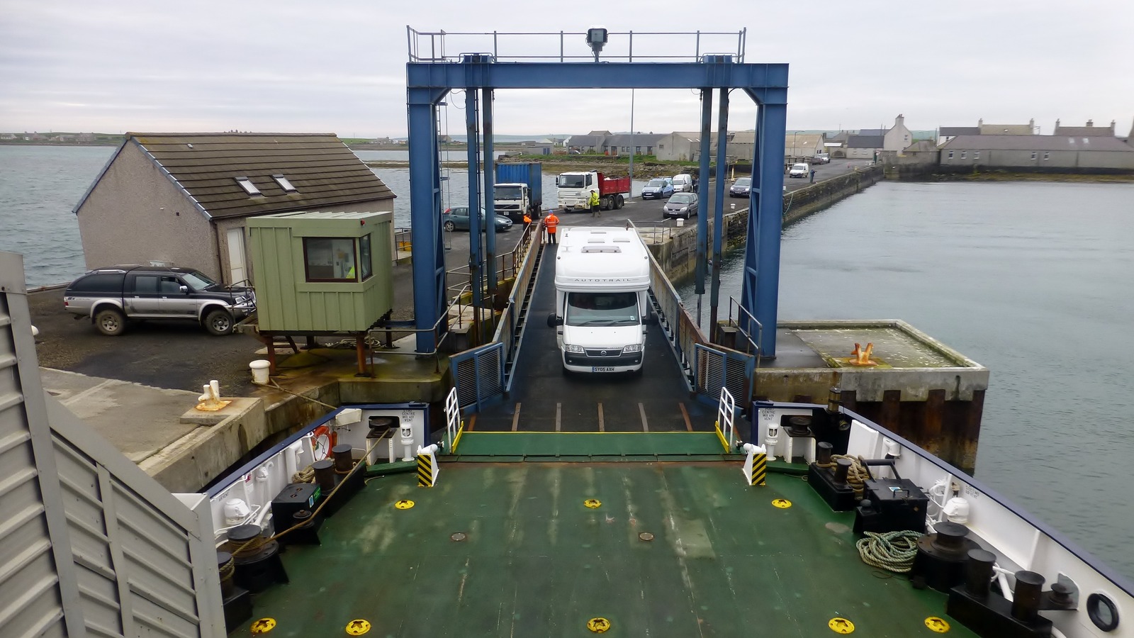 The green vehicle deck and stern ramp of an Orkney Ferries vessel at the pier on Stronsay, with a white motorhome driving aboard and lorries and cars waiting on the quayside in front of the village