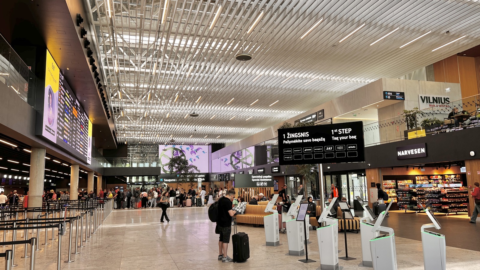 Interior of the Vilnius Airport departure terminal in June 2026, with bag-drop kiosks, a flight board, and a Vilnius bar sign.