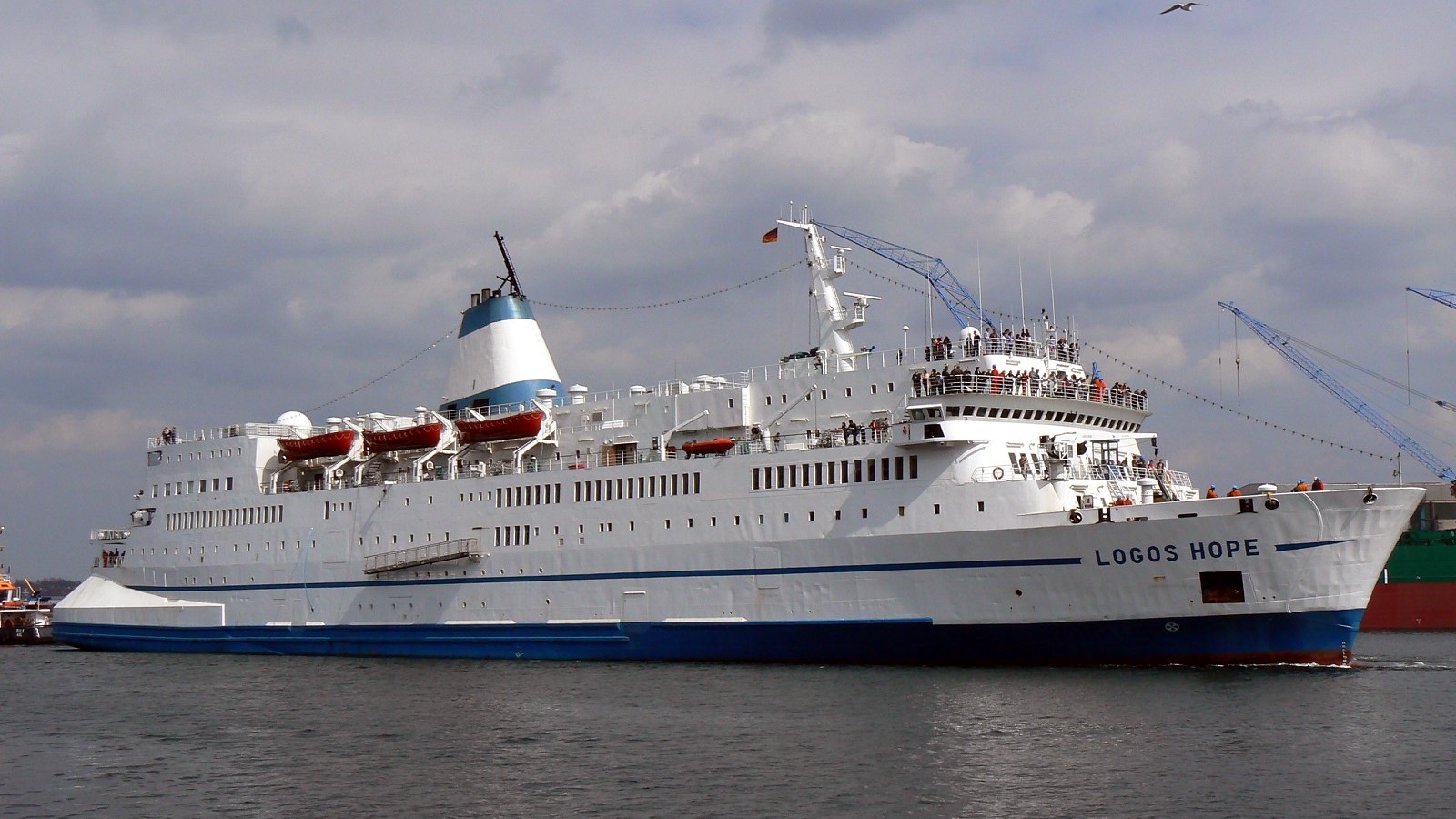 White ocean-going ship Logos Hope with a blue hull stripe and the name painted on the bow, photographed in a harbor