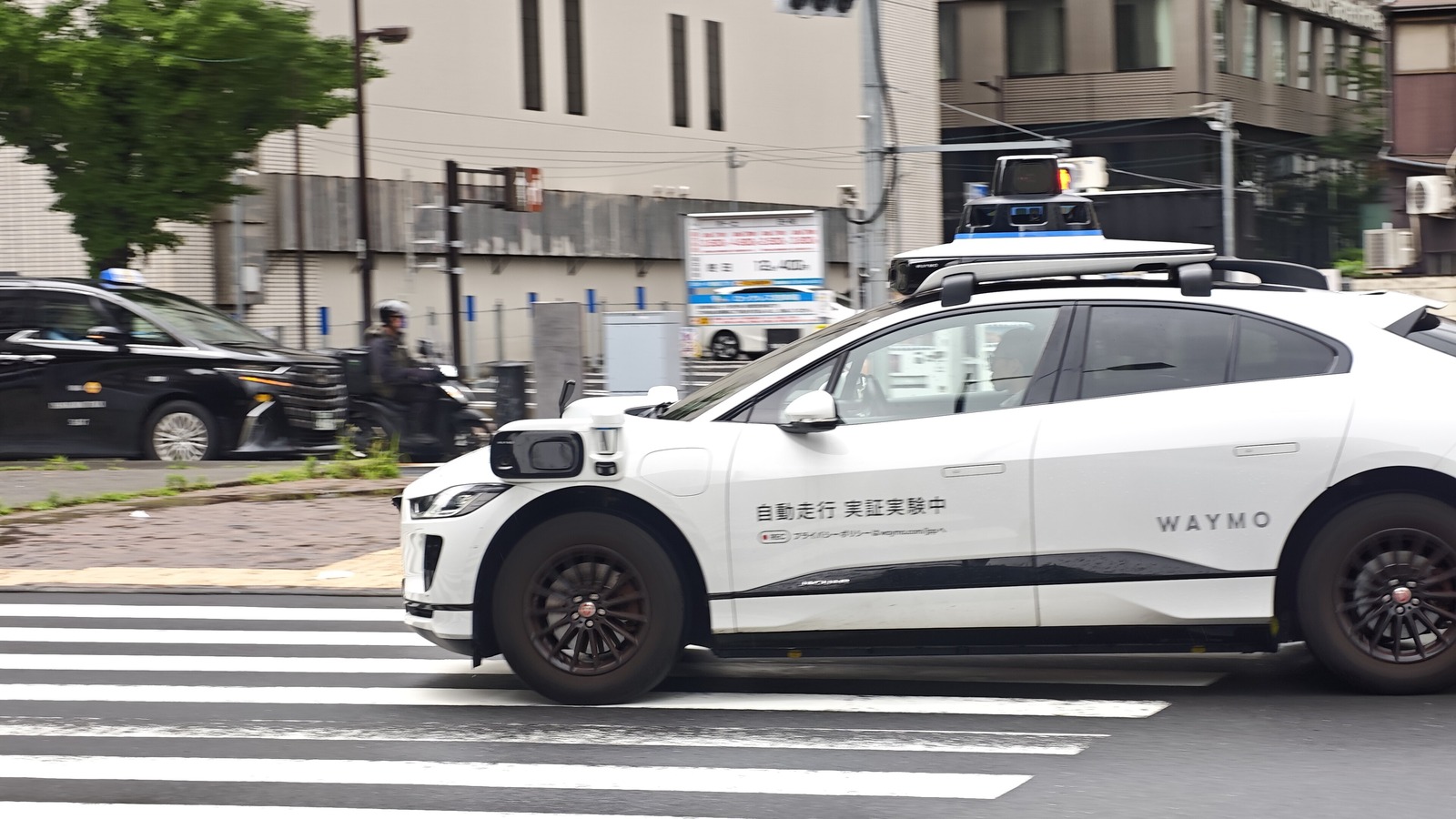 A white Waymo Jaguar test car with roof sensors and a Japanese autonomous driving trial sign, crossing a street in Tokyo in June 2026.