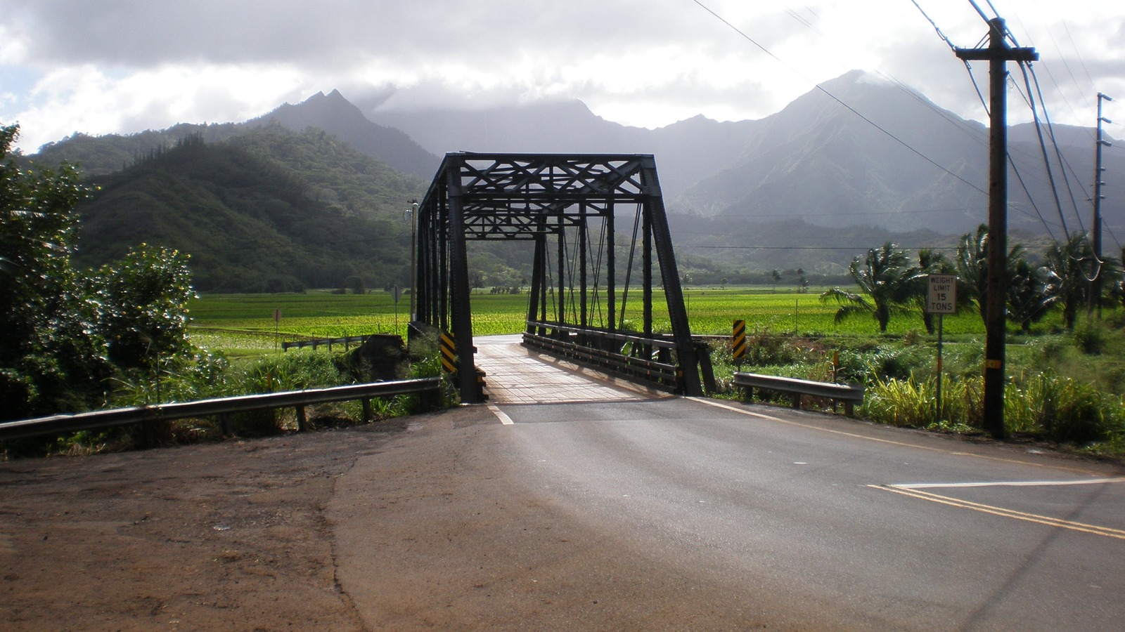 The one-lane steel Hanalei Bridge on Kuhio Highway with utility poles, green taro fields and mountains behind it