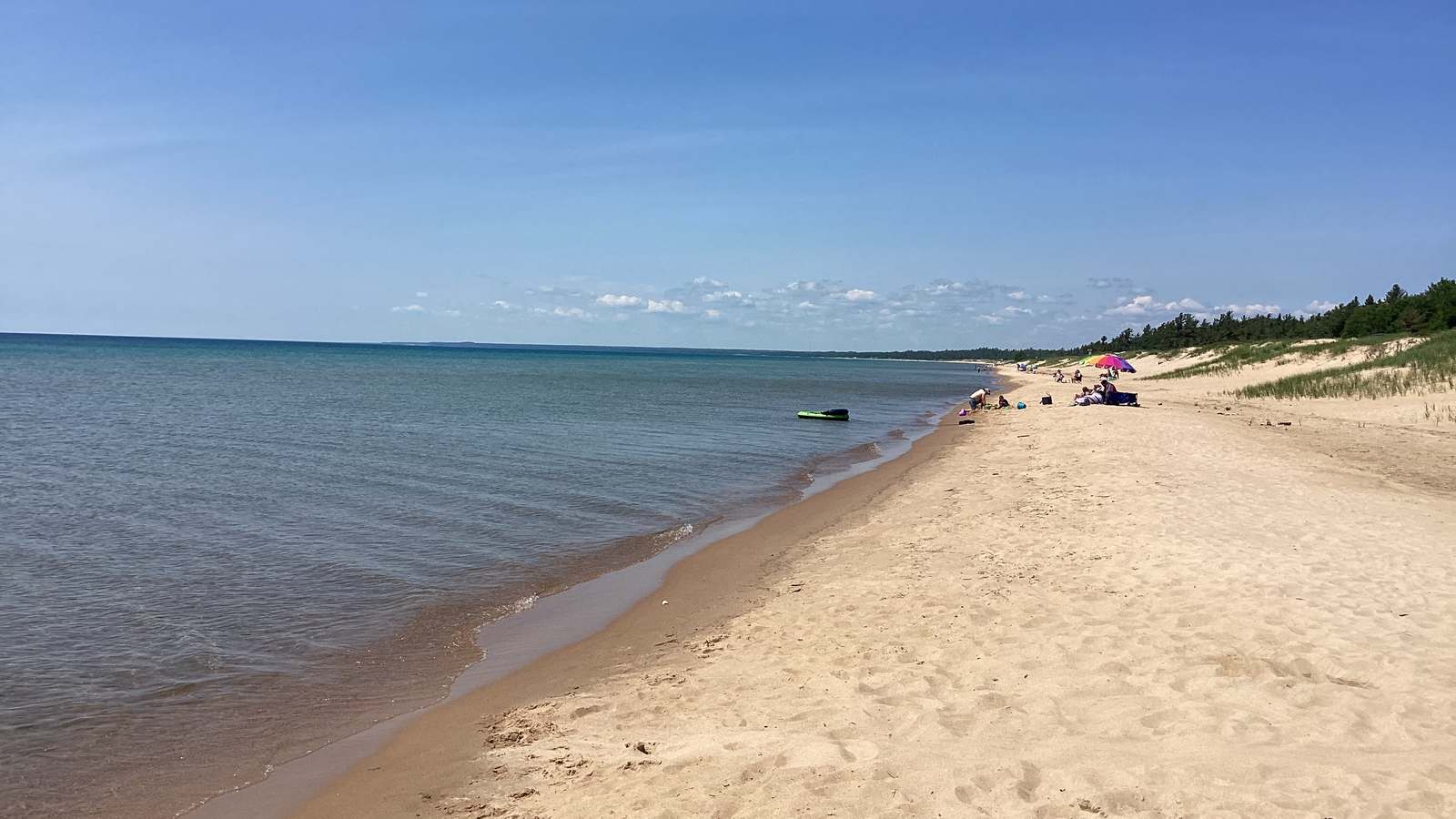 Sand Point Beach on Lake Superior at Pictured Rocks National Lakeshore, with clear shallow water, a wide sand beach and a wooded dune line under a blue sky.