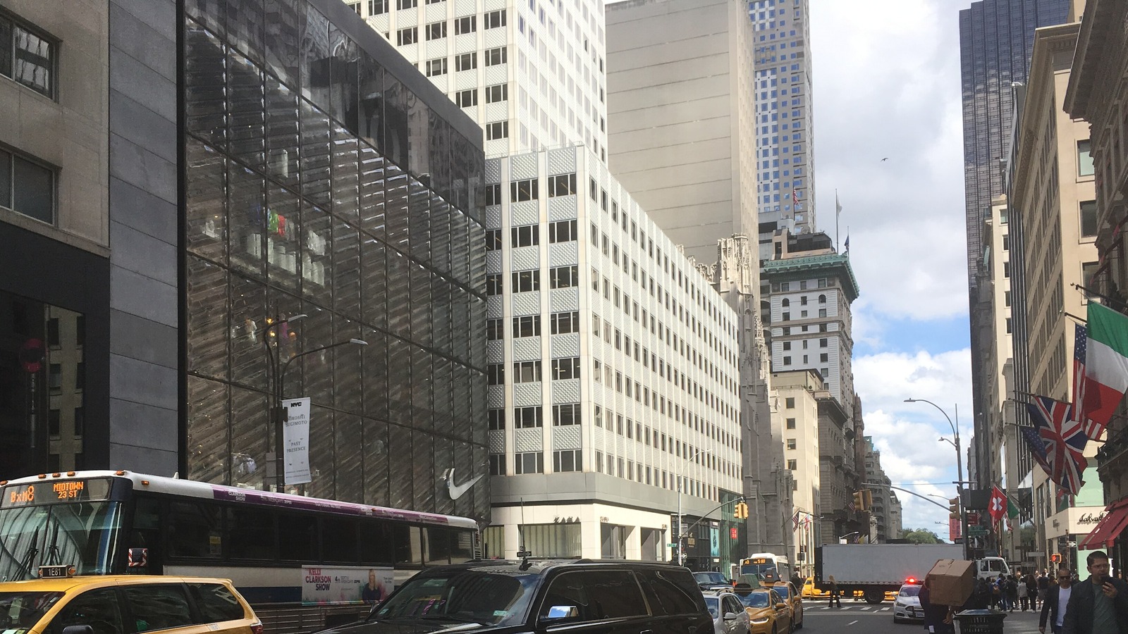 Fifth Avenue in Manhattan looking north from 51st Street on an ordinary weekday, with taxis, a city bus and a box truck filling both lanes between tall office buildings, traffic signals and street lights overhead, a row of national flags projecting from a building on the right and a cloudy sky above