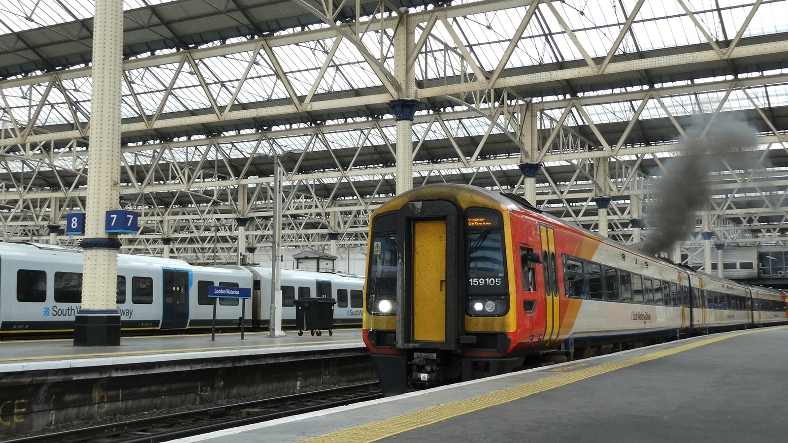 A class 159 diesel unit numbered 159105 standing under the trainshed at London Waterloo with its destination display reading Exeter St Davids, a South Western Railway suburban unit at the adjacent platform and the London Waterloo platform nameboard in the background