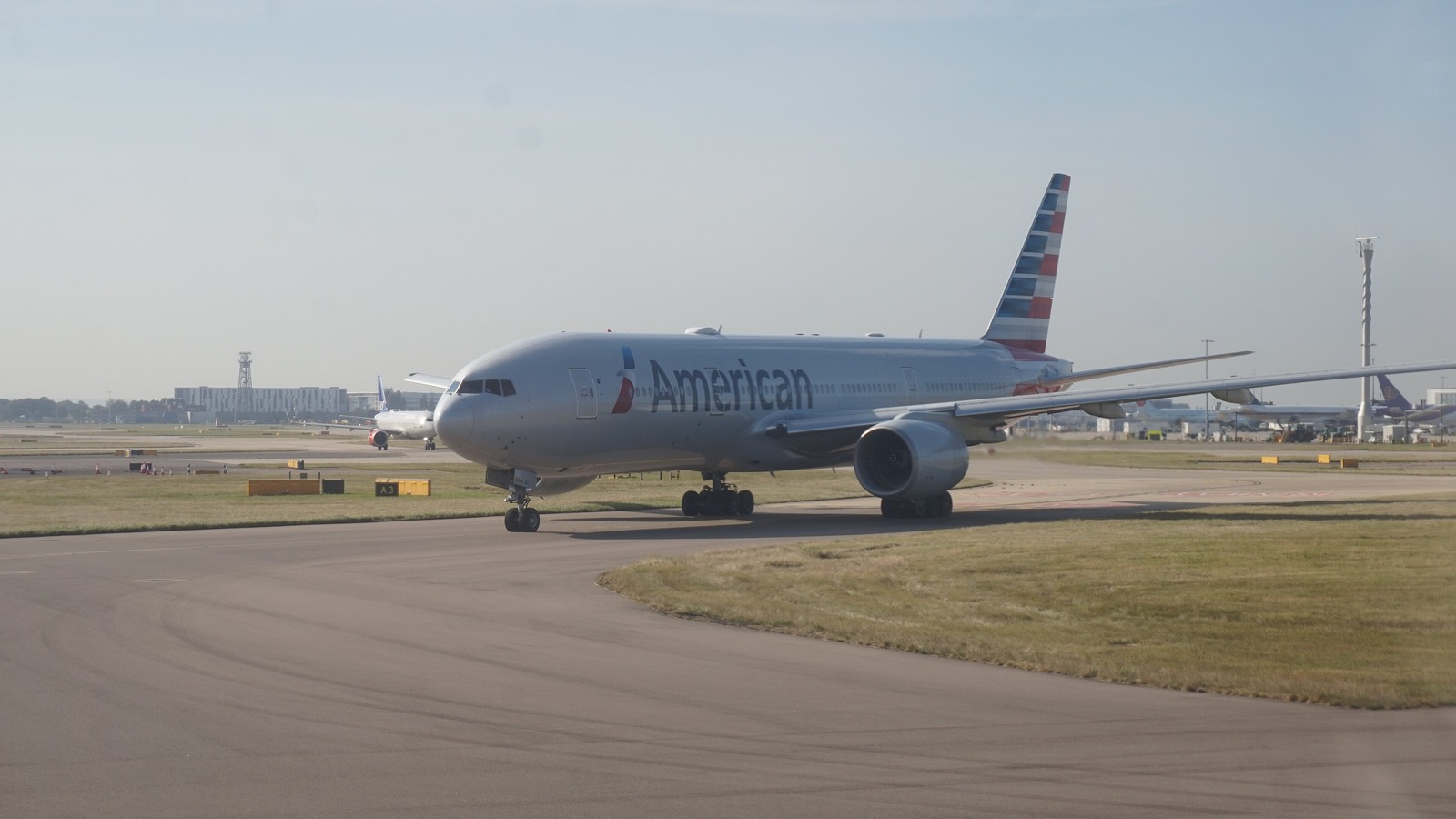 An American Airlines widebody taxiing toward runway 27R at London Heathrow on a hazy morning, with other aircraft and terminal buildings along the far side of the airfield.