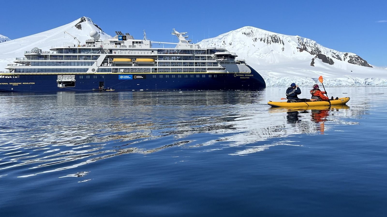 Two kayakers paddle in calm Antarctic water in front of the expedition ship National Geographic Resolution, its Lindblad Expeditions and National Geographic wordmarks visible on the dark blue hull, with snow covered peaks behind