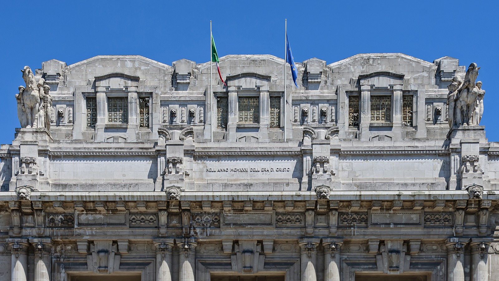 Travelers with luggage cross the plaza in front of the entrance portico at Milano Centrale railway station under a clear sky.