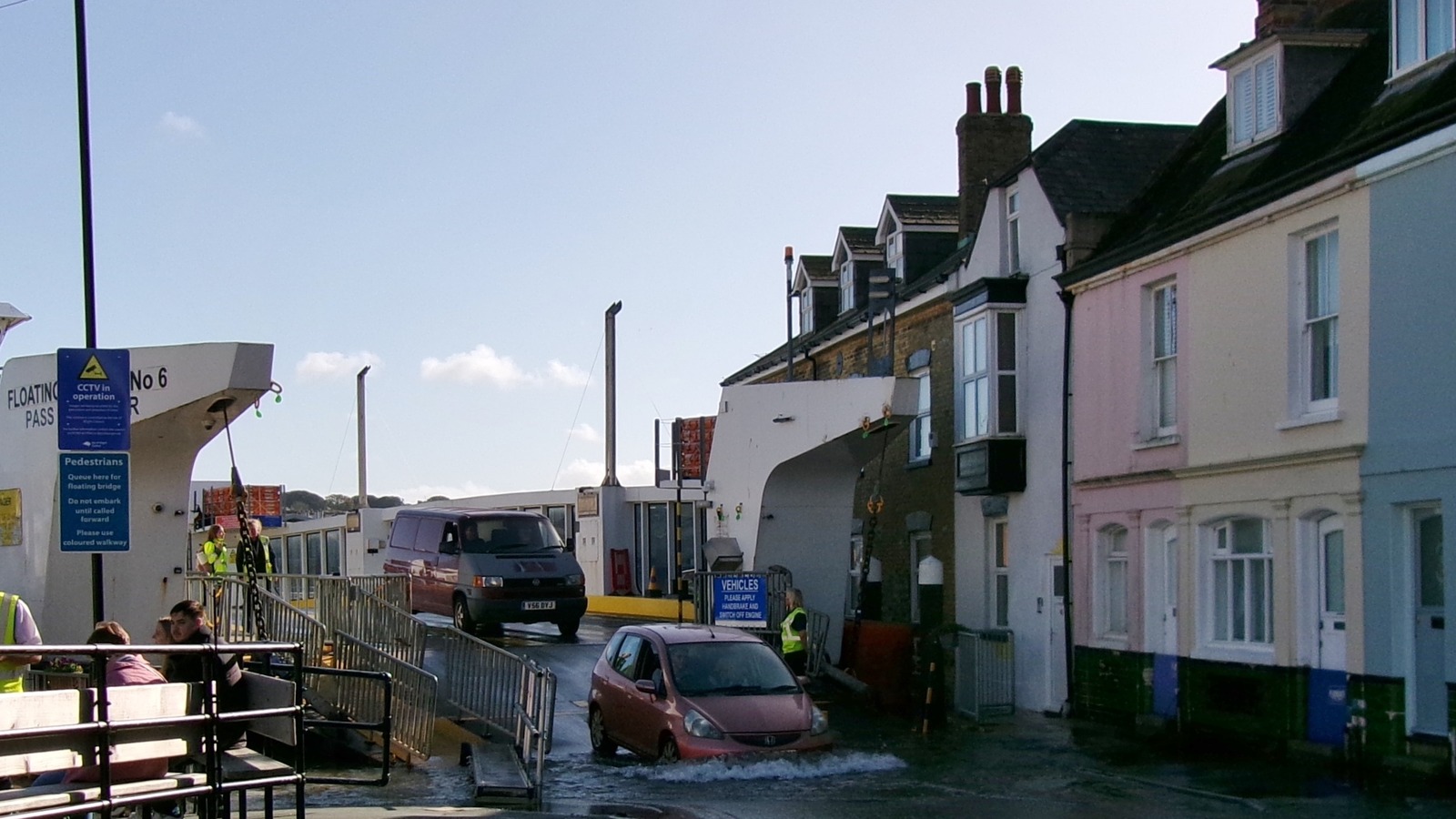Cowes Floating Bridge No. 6 at the Isle of Wight slipway, with vehicles leaving the chain ferry beside riverside houses.