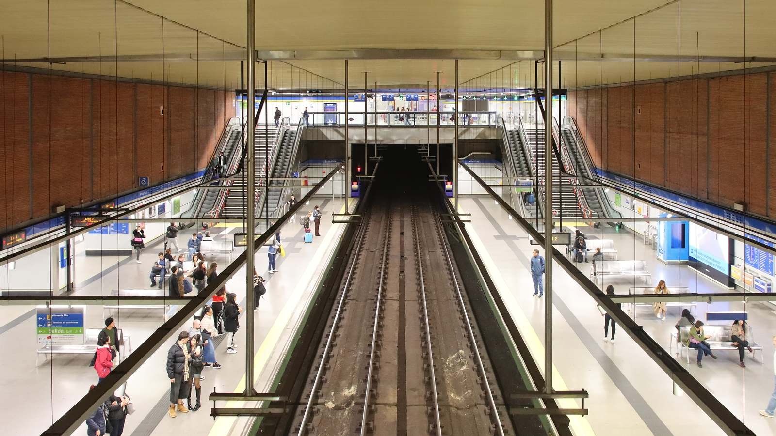 Moncloa metro station in Madrid seen from above, with two platforms, escalators and riders waiting beside the tracks.
